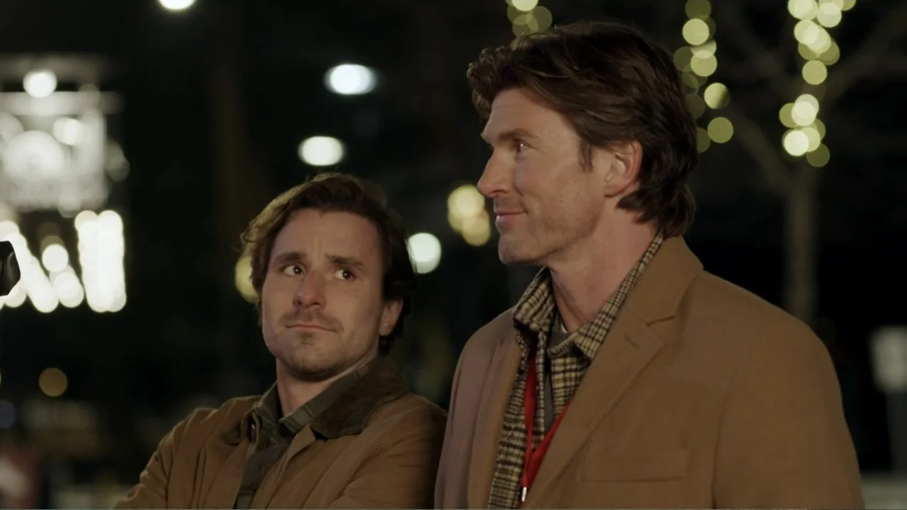 Peppermint and Postcards (2023) movie still: medium — Two men stand outdoors at night in front of blurred circular glowing bokeh lights. They stand…