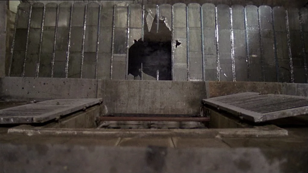 The Tenant (1976) movie still: establishing — An industrial utility area features a broken glass pane in a paneled wall. Two heavy metal grate…