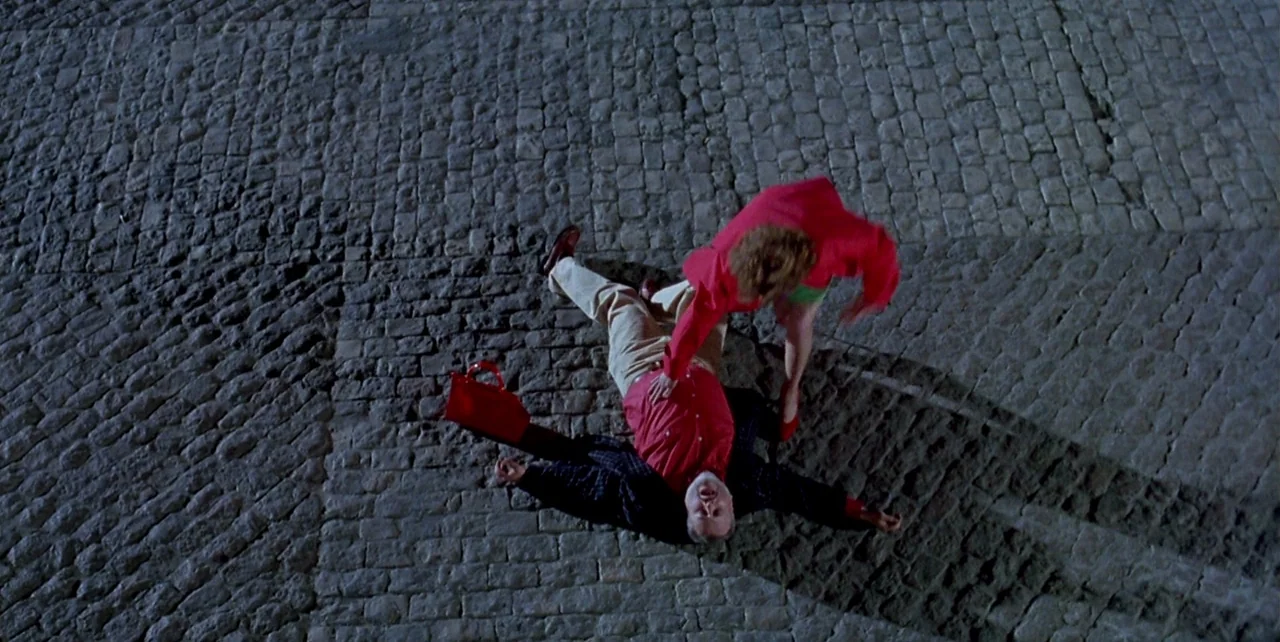 The Flower of My Secret (1995) movie still: wide — A dark cobblestone ground is illuminated by a single harsh overhead light. A woman in a red…
