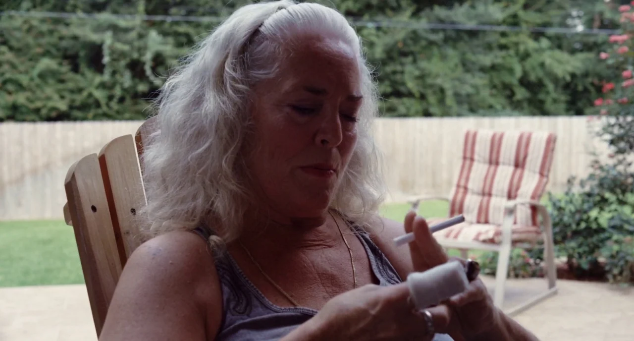 Krisha (2015) movie still: medium — An outdoor backyard area features a wooden patio chair and lawn furniture. An older woman with white hair holds…