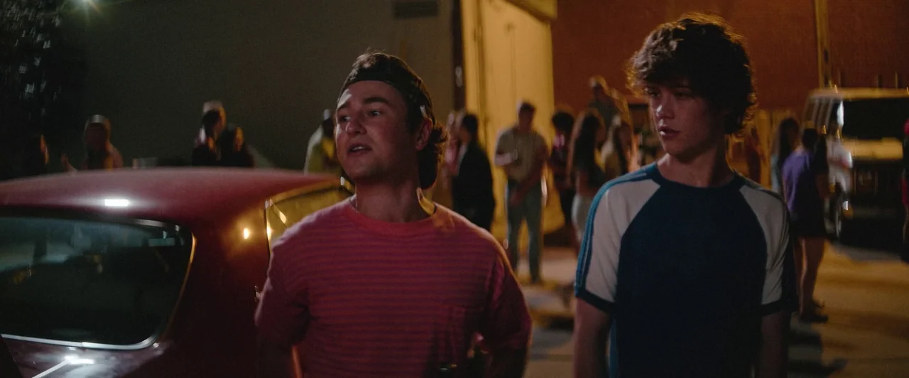 Snack Shack (2024) movie still: medium — A group of people gathers in a dark, paved area at night. Two young men stand in the foreground talking…