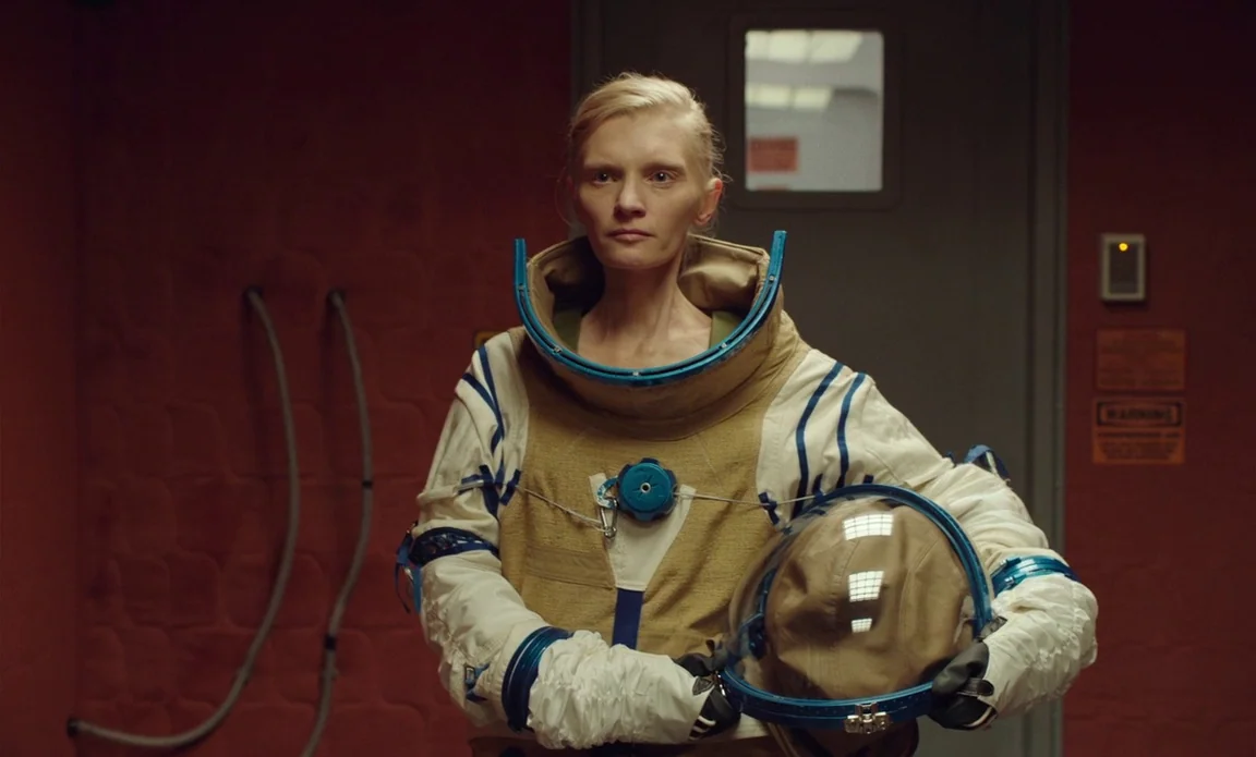 High Life (2018) movie still: medium — A red-walled room features a gray metallic door with a rectangular observation window. A blonde woman in a…