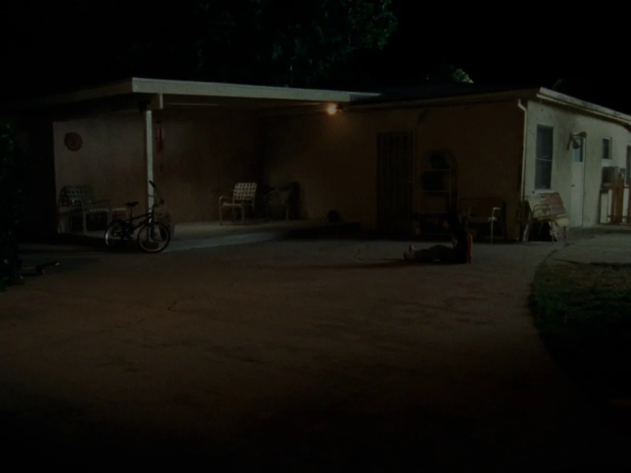 Mid90s (2018) movie still: wide — A dark, single-story house features a concrete driveway and an attached covered carport area. A lone individual…