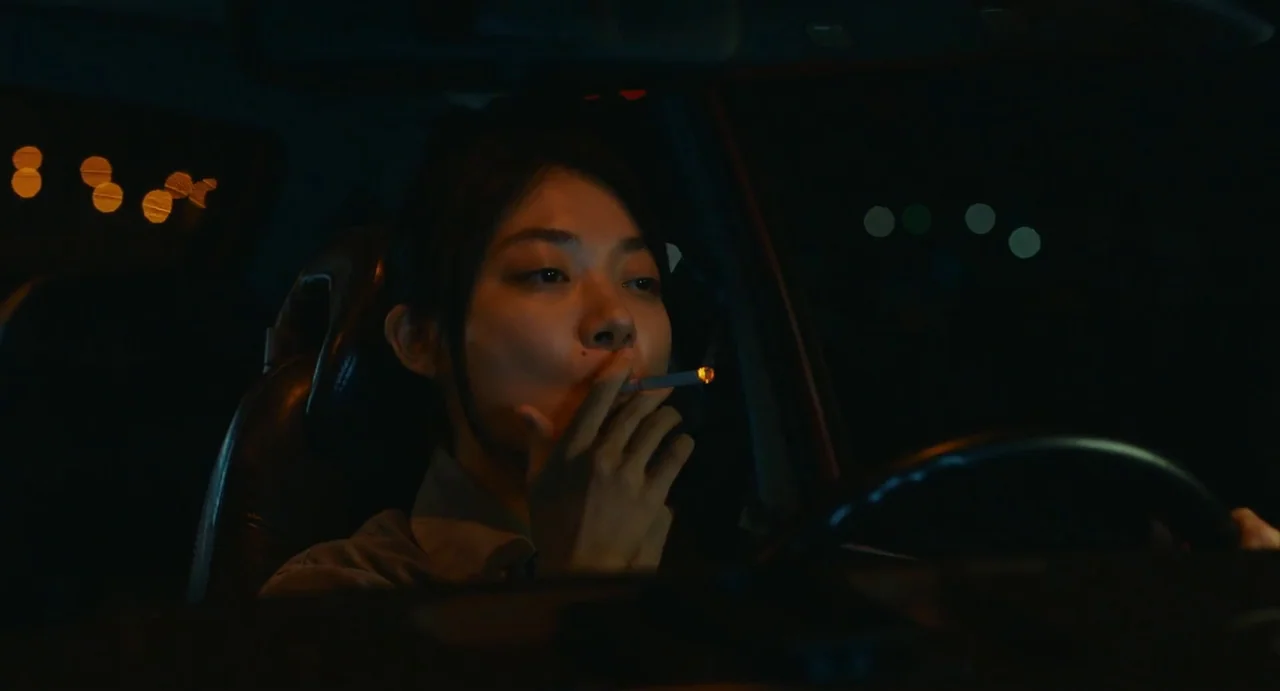Drive My Car (2021) movie still: closeup — A dark car interior at night with the subject sitting in the driver's seat. The woman holds a lit…