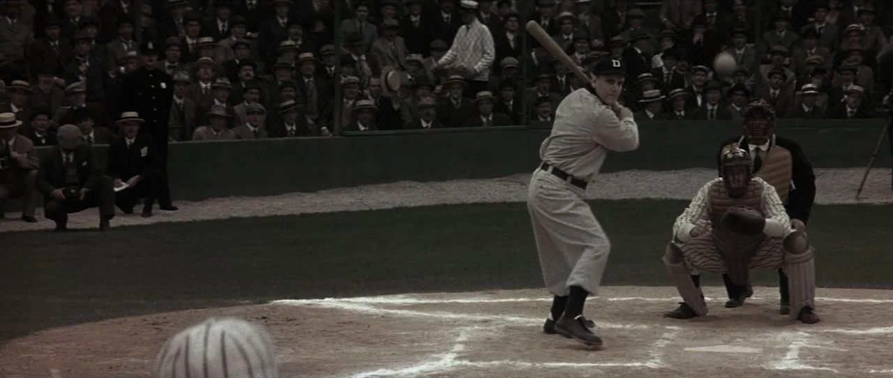 Cobb (1994) movie still: medium — A historic baseball park features a dirt home plate surrounded by a dense crowd. A batter in vintage grey uniform…