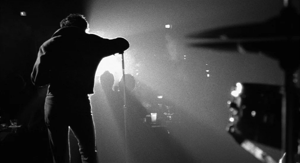 Lenny (1974) movie still: medium — A dark, smoky room with tables and glasses visible in the mid-ground. A man stands on a stage leaning against a…