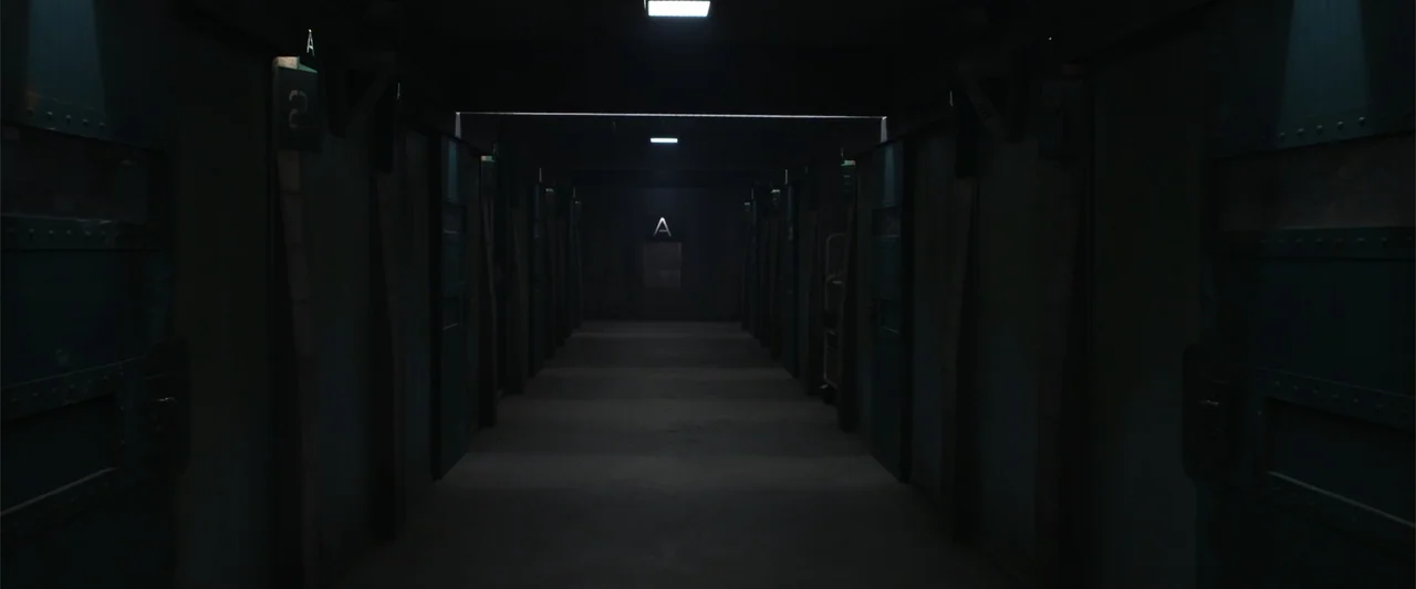 Black Site (2022) movie still: establishing — A dark industrial corridor is lined with heavy metal doors marked with numeric labels. The long narrow…