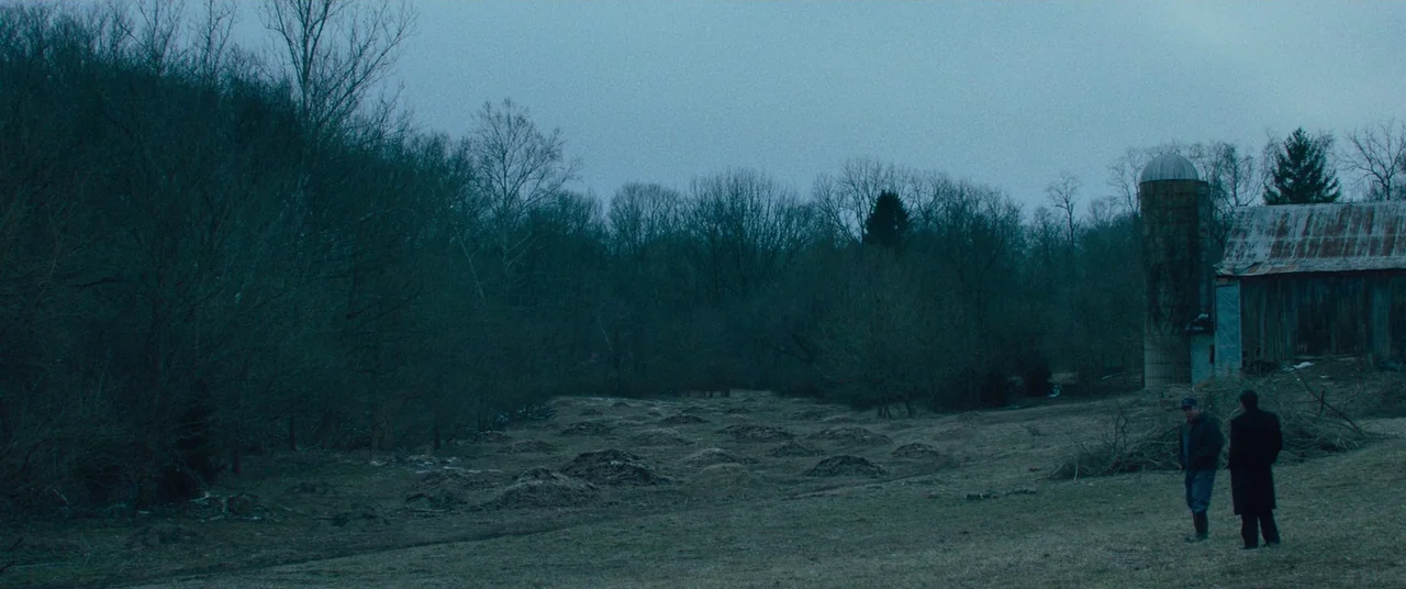 Dark Waters (2019) movie still: wide — A rural expanse features rows of fresh dirt mounds leading toward a weathered barn. Two men stand together in…