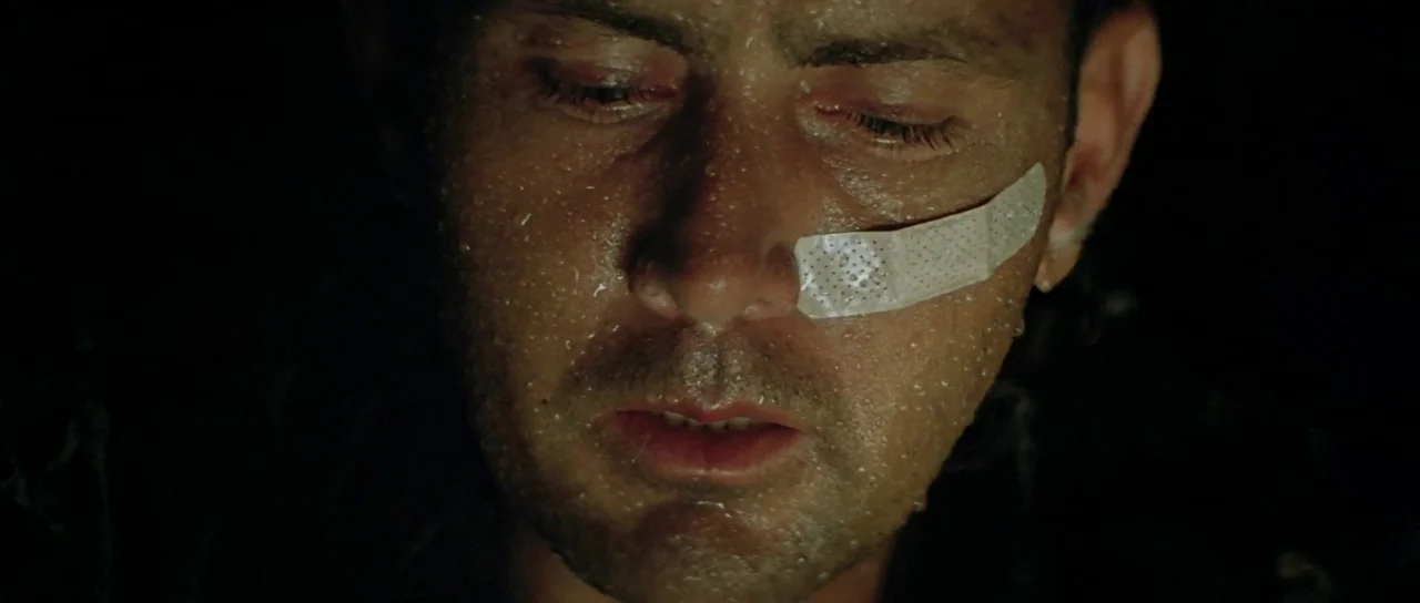 Apocalypse Now (1979) movie still: closeup — A close-up of a man in deep shadow against an indistinct dark background. The man has beads of sweat on…