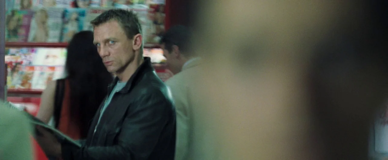 Casino Royale (2006) movie still: medium — A man stands in front of a shelf stocked with various colorful magazines. He turns his head to look…