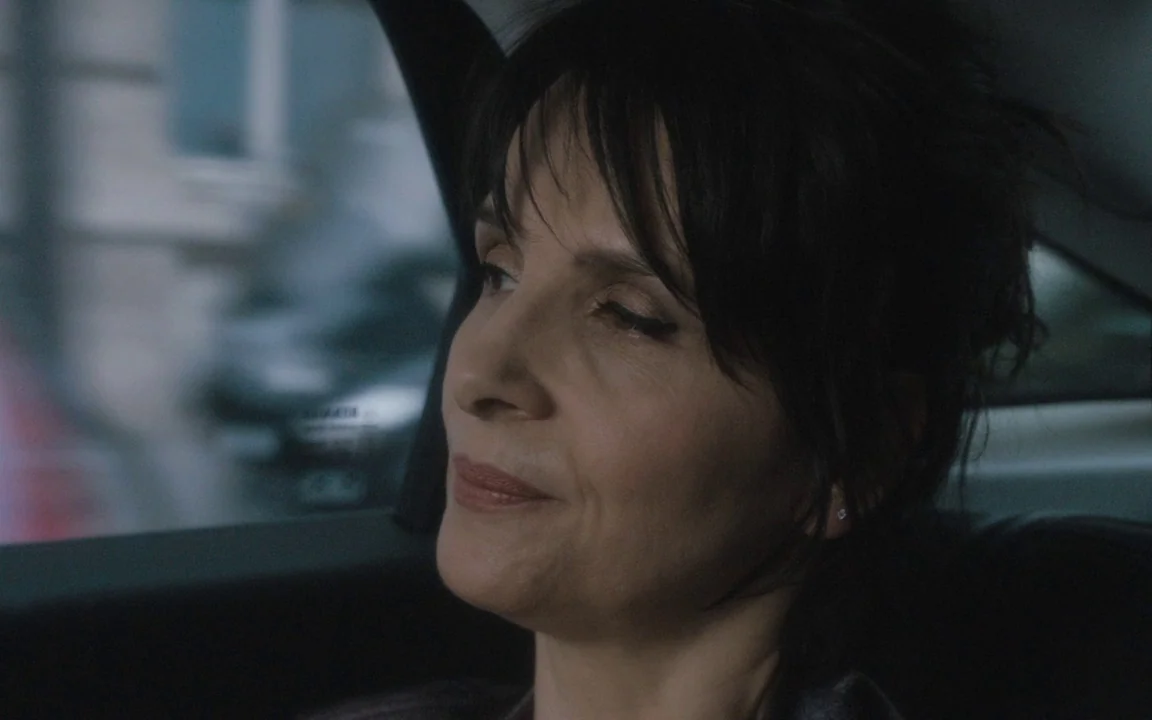 Let The Sunshine In (2017) movie still: closeup — A woman sits in the back seat of a moving vehicle. She gazes out the window as the city blur…