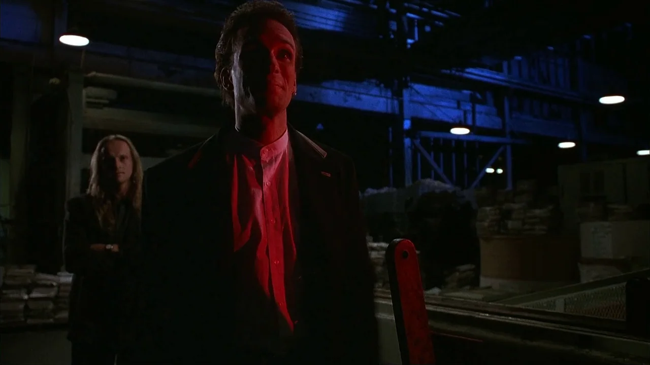 The Mask (1994) movie still: medium — An industrial facility at night with dimly lit rafters and stacks of stored goods. A man with short dark hair…