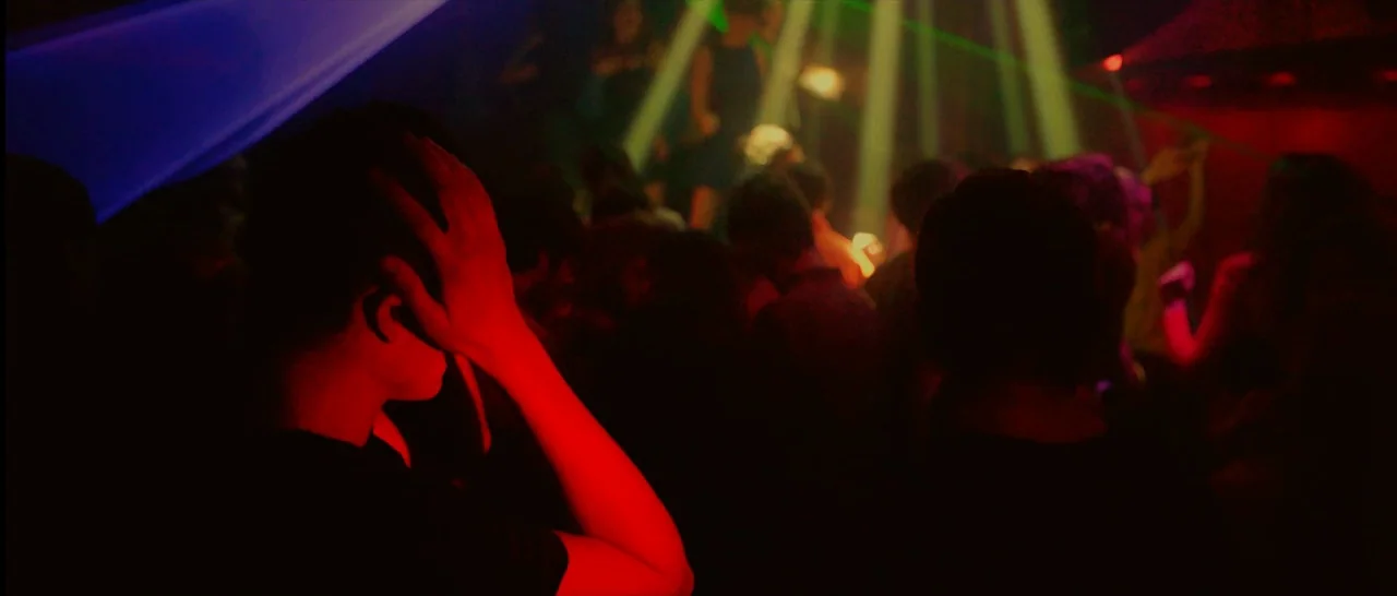 Love (2015) movie still: medium — A dark, crowded room is filled with atmospheric smoke and intersecting beams of light. A person stands in the…