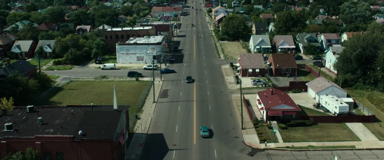 Don’t Breathe (2016) movie still: establishing — An aerial view shows a wide suburban road intersecting a neighborhood of houses and buildings. Two…