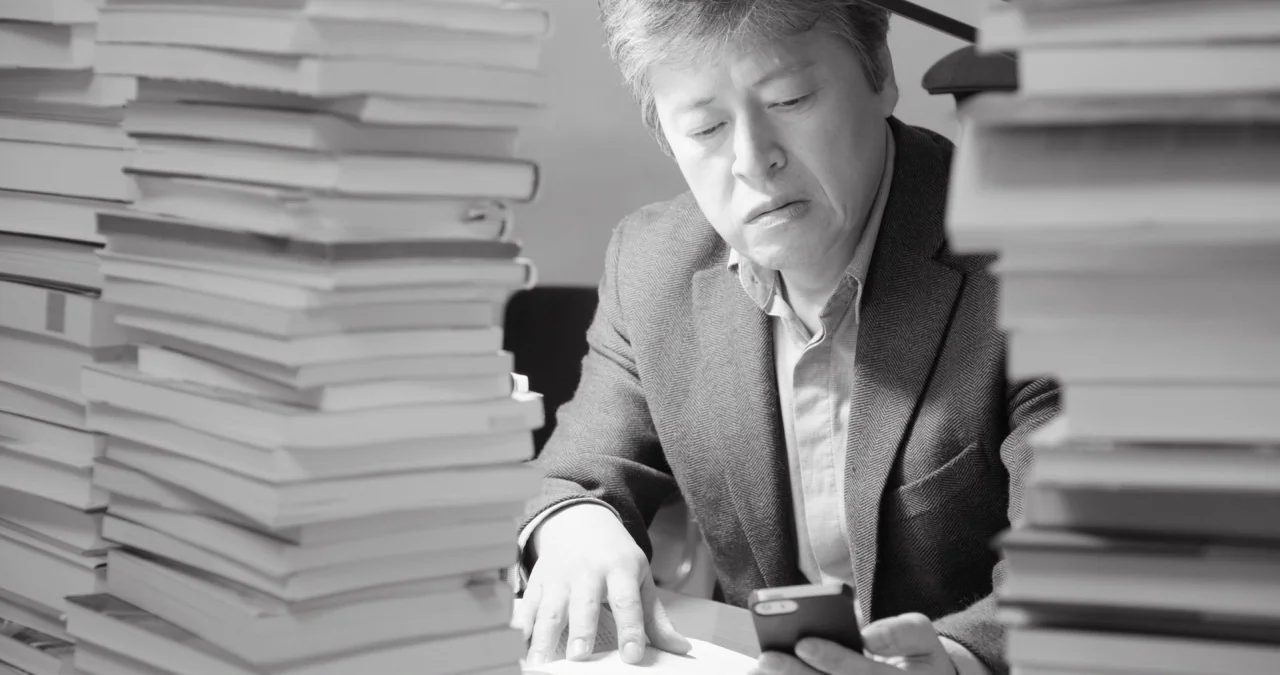 The Day After movie still: medium — A workspace features large stacks of books framing a seated man on both sides. The man sits at a desk while…