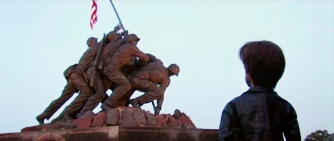 Team America: World Police movie still: medium — A bronze sculpture depicting the Iwo Jima flag raising stands atop a stone base. A young boy seen…