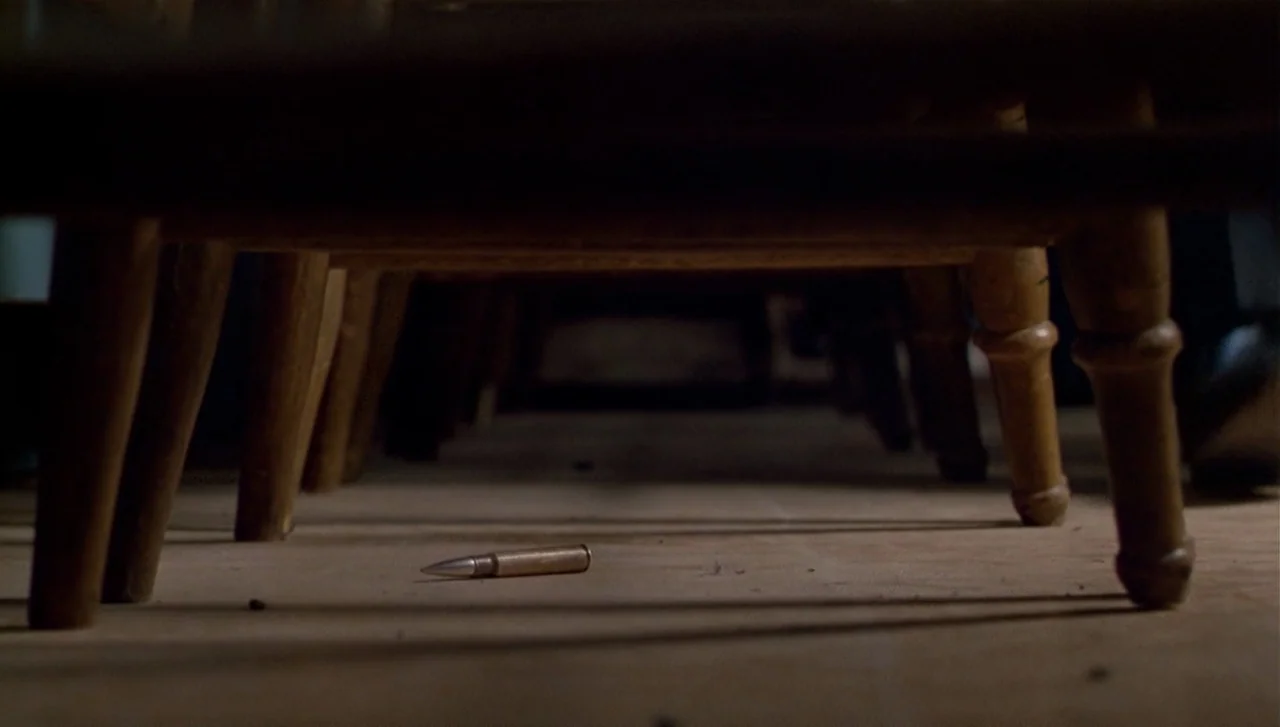 The Dead Zone (1983) movie still: closeup — A low-angle view shows the space beneath several rows of wooden chairs. A single metal rifle cartridge…