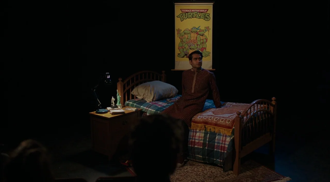The Big Sick (2017) movie still: wide — A man sits on a bed placed on a dark, minimalist theatrical stage. A Teenage Mutant Ninja Turtles poster…
