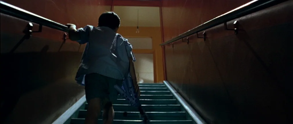 Mortal Transfer (2001) movie still: fullbody — A narrow stairwell features orange walls and painted teal wooden steps. A young boy walks upward…