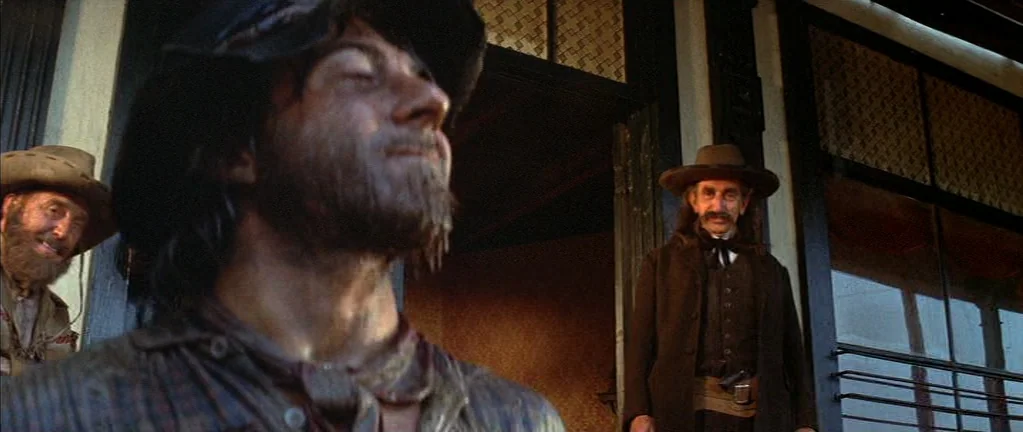 Little Big Man (1970) movie still: medium — A wooden boardwalk in front of a storefront with glass windows and dark wood. A disheveled man stands in…