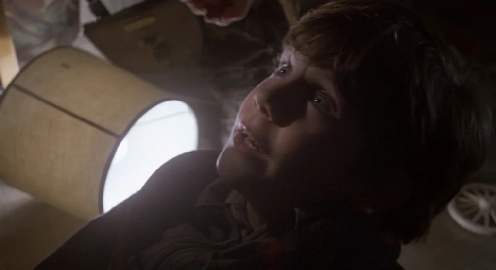Time Bandits (1981) movie still: closeup — A dark, cluttered space contains a young boy looking up at a bright light. The boy is positioned near a…