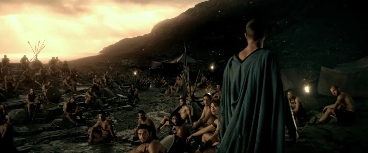 300: Rise of an Empire (2014) movie still: medium — A dark rocky shoreline serves as a military camp at the edge of the water. A man in a blue cape…