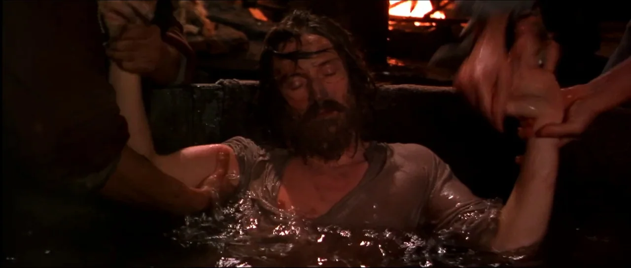 Ravenous (1999) movie still: medium — A dark stone cell contains a large water vessel illuminated by a distant fire. A man with matted hair and a…