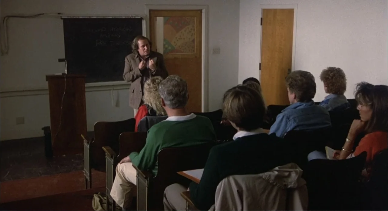 Back to School (1986) movie still: medium — A plain classroom features a black chalkboard, a wooden lectern, and rows of seats. A middle-aged man…
