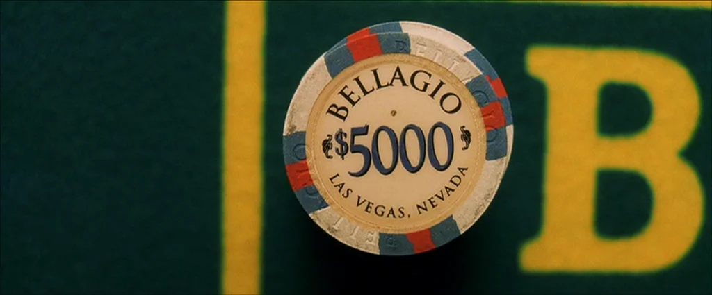 Ocean’s Eleven (2001) movie still: closeup — A top-down view shows a single casino chip placed on a green felt surface. The chip features text…