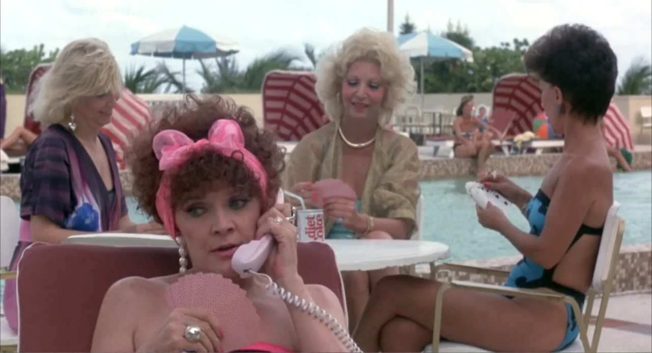 Making Mr. Right (1987) movie still: medium — A pool deck with several women in 1980s swimwear sitting around white plastic tables. One woman in the…