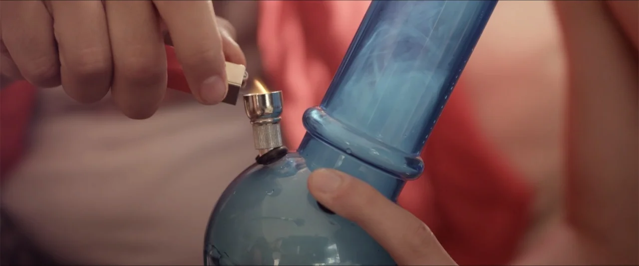 High Five (2004) movie still: closeup — A close view shows a person's hands holding a blue water pipe. One hand holds a small red lighter to the…