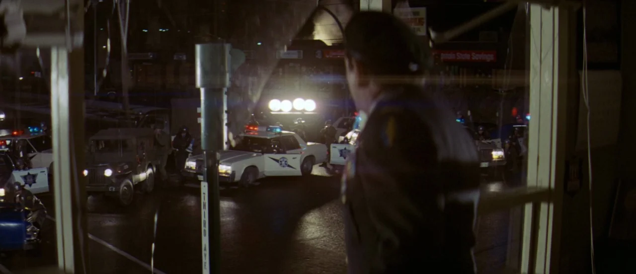 First Blood (1982) movie still: medium — A dark, windowed room overlooks a rainy urban street occupied by police vehicles. A man in uniform stands…