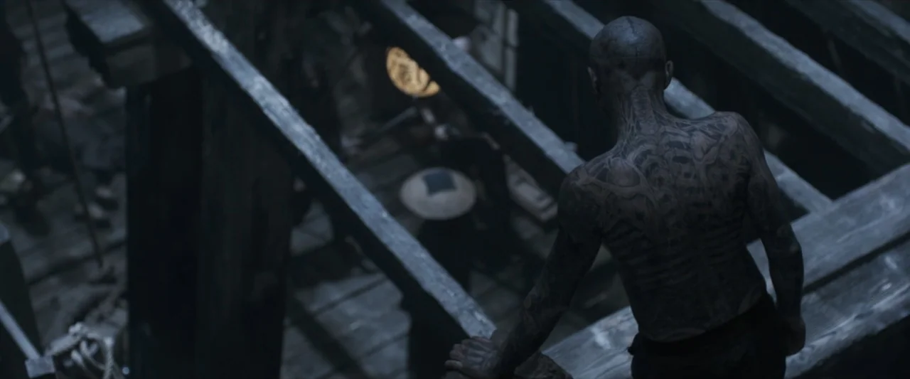 47 Ronin (2013) movie still: medium — A dark wooden structure features heavy exposed beams and a view to a lower level. A tattooed man stands on a…