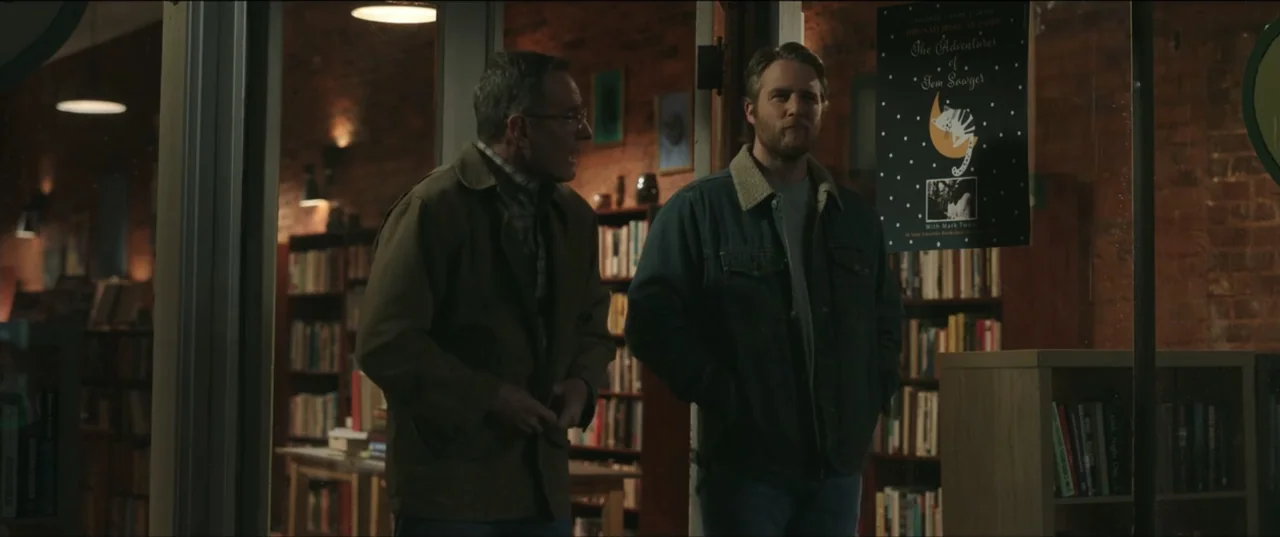 Jerry & Marge Go Large (2022) movie still: medium — A dimly lit bookstore interior features rows of shelves filled with numerous books. Two men in…
