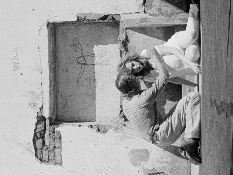 Tabu (2012) movie still: medium — A weathered brick wall with peeling plaster and a simple chalk stick figure drawing. A man and a woman sit…