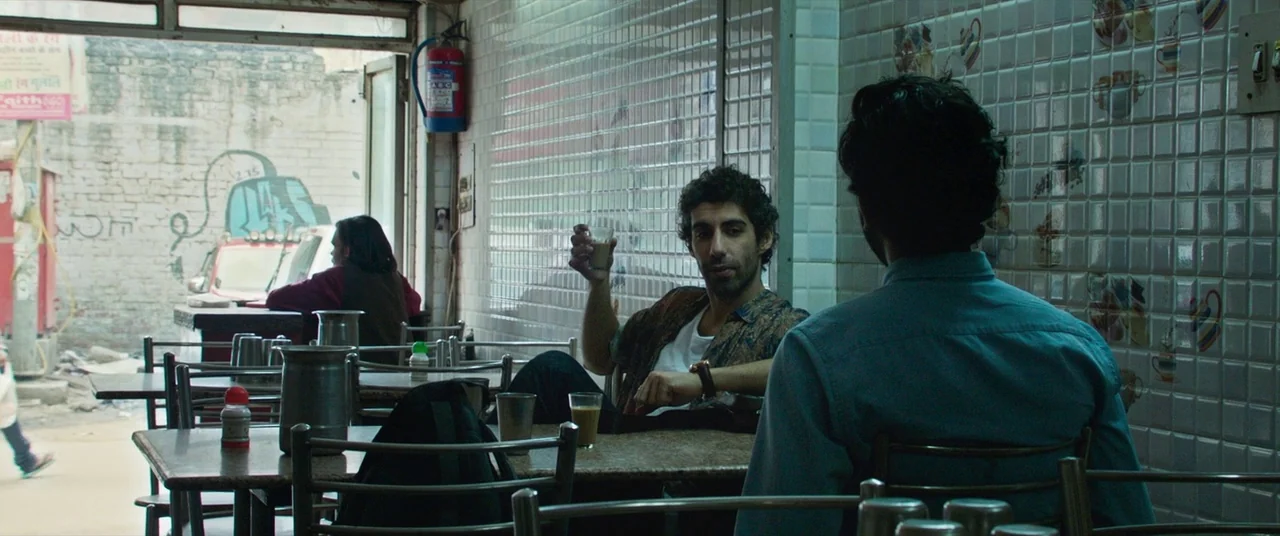 The Wedding Guest (2018) movie still: medium — A utilitarian street-side tea stall features white tiled walls and metal tables. Two men sit at…