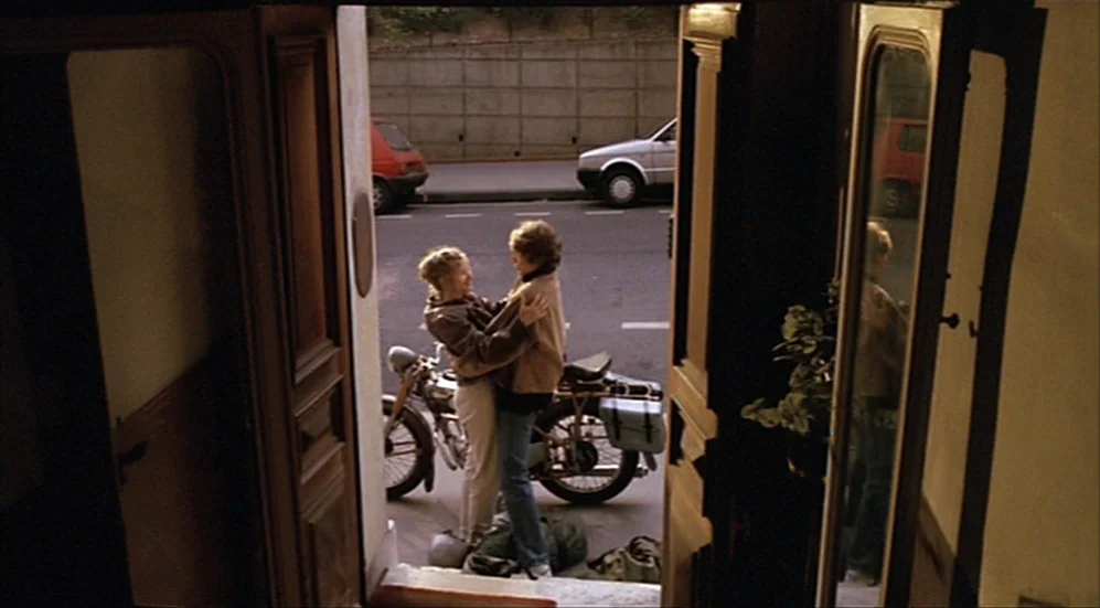 Roselyne and the Lions (1989) movie still: fullbody — A sidewalk with parked motorcycles and a car visible in the background. Two young women stand…