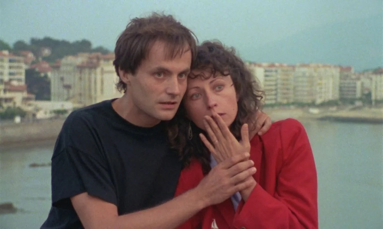 The Green Ray (1986) movie still: medium — A coastal promenade features distant apartment blocks along a curved shoreline in the background. A man…