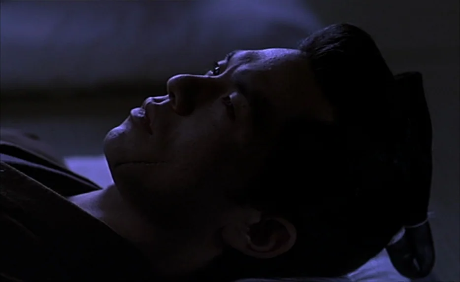 Gohatto (1999) movie still: closeup — A man lies on a pillow in a dimly lit, dark blue environment. He gazes upward with eyes open in the quiet space.