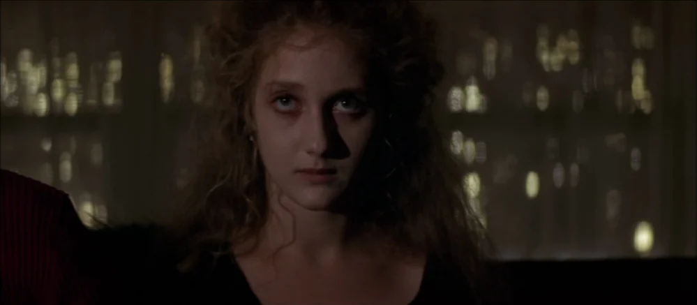 Carnal Knowledge (1971) movie still: closeup — A dimly lit room features a woman centered against a dark, bokeh-filled backdrop. The subject gazes…