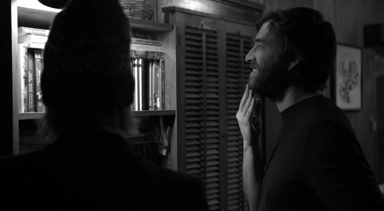 Blue Jay (2016) movie still: medium — A room with wooden shelving filled with books and a louvered cabinet door. A bearded man looks toward the…