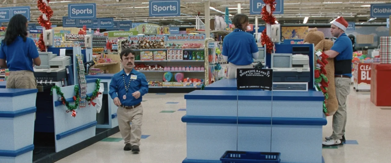 Roofman (2025) movie still: medium — A retail store checkout area features blue counters and overhead signage for sports goods. A man of short…