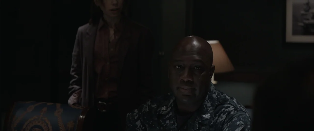 Godzilla (2014) movie still: medium — A dimly lit room features a man in naval uniform sitting in the foreground. A woman stands behind him in the…