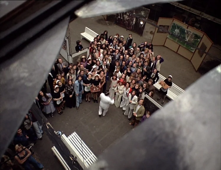 The Arrival of Joachim Stiller (1976) movie still: establishing — A large paved courtyard is surrounded by buildings and filled with a dense crowd.…