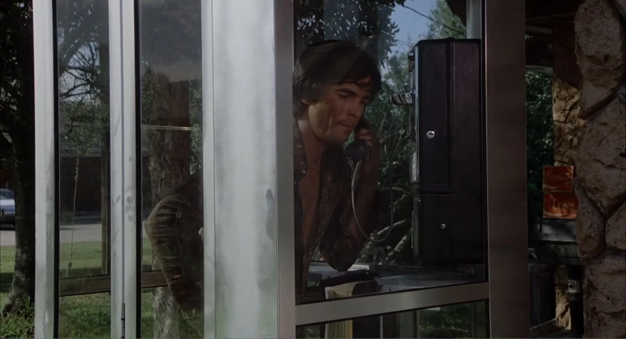 Empire of the Ants (1977) movie still: medium — A glass telephone booth sits outdoors near a stone wall and green trees. A young man stands inside…