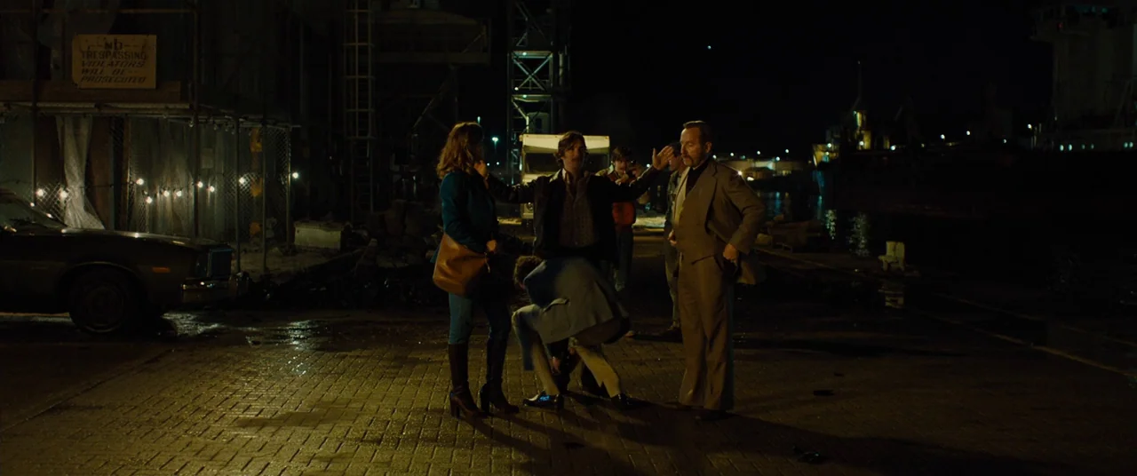 Free Fire (2016) movie still: wide — A dark industrial shipping area at night with wet pavement and construction fencing. A small group of…