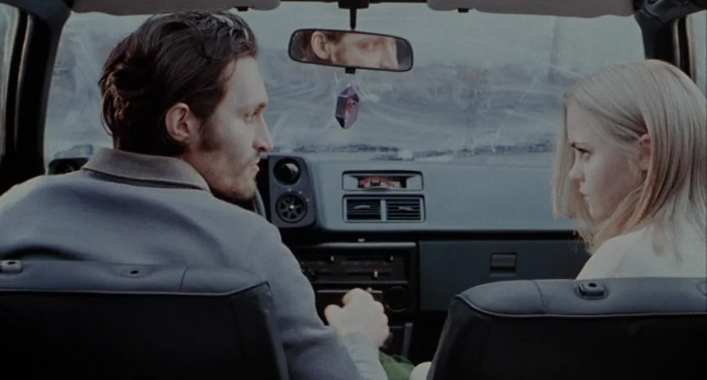 Buffalo ’66 (1998) movie still: medium — The front seats of a car with a black dashboard and rear-view mirror. A man looks toward a woman sitting in…