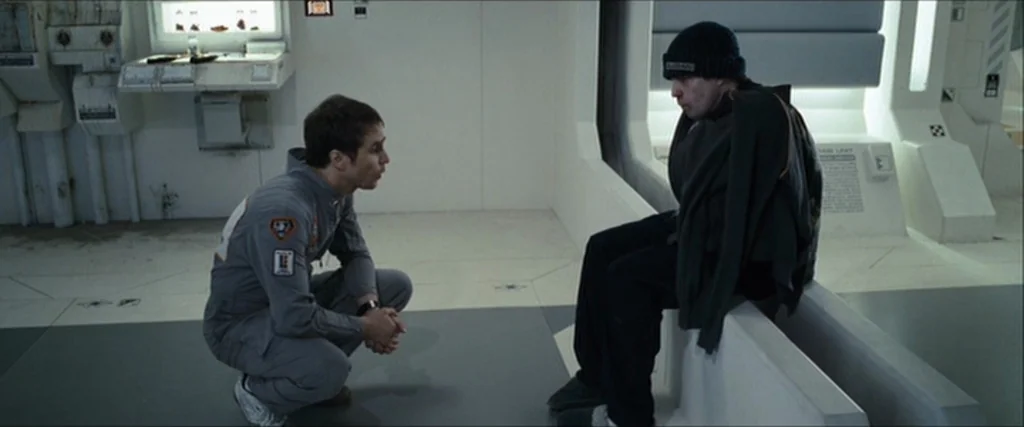 Moon (2009) movie still: medium — A sterile, minimalist white spaceship interior with modular panels and white flooring. Two men interact in the…