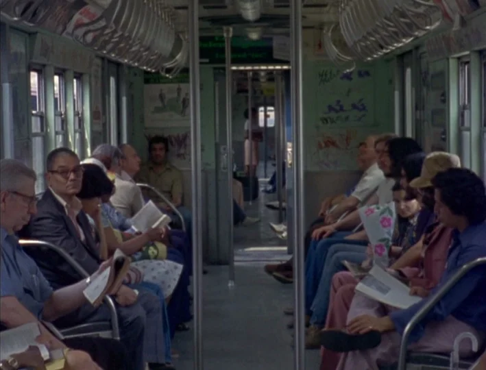 News From Home (1977) movie still: wide — An interior of a 1970s New York City subway car with green-painted walls and graffiti. Passengers sit in…