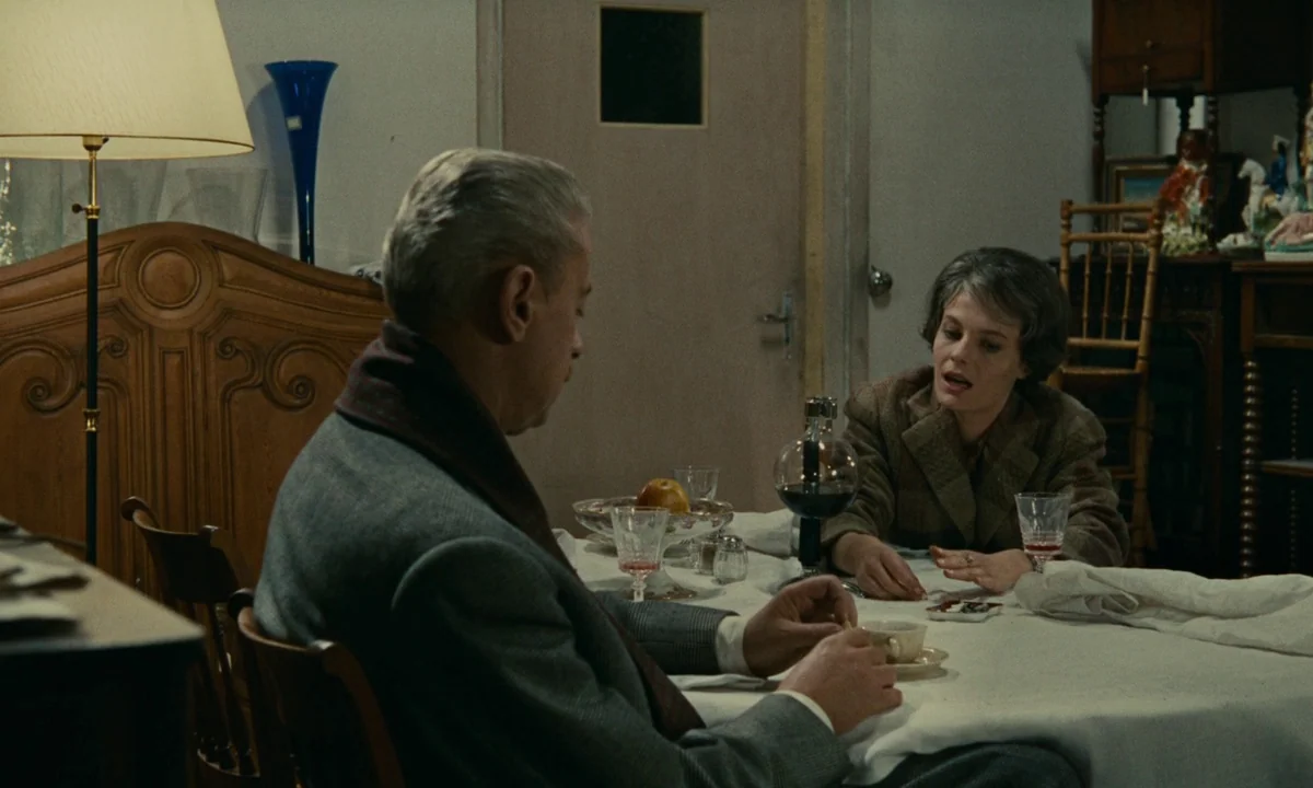 Muriel (1963) movie still: medium — A man and woman sit across from each other at a covered dining table. The room features dark wood furniture, a…